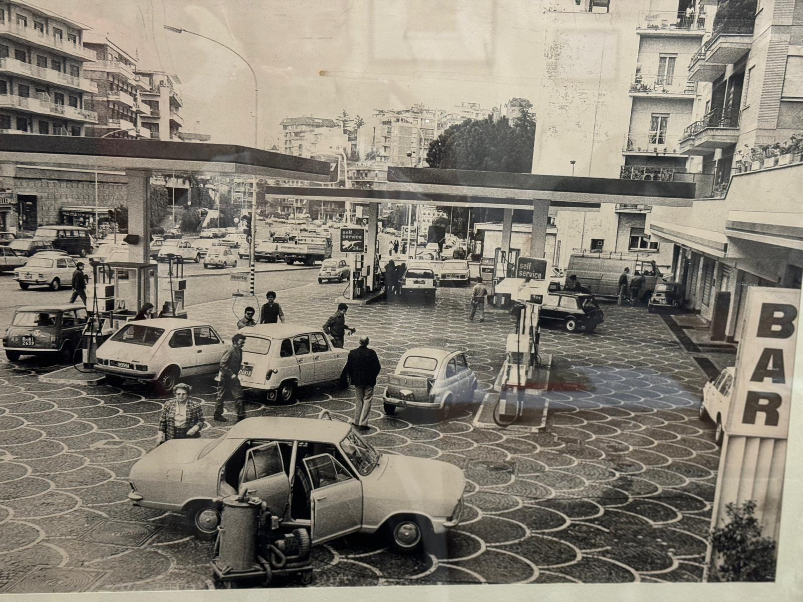 Una scena d'epoca si svolge presso la vivace stazione di servizio, dove il gestore stazione di servizio supervisiona il rifornimento di più auto e l'interazione delle persone. Nelle vicinanze, un cartello "BAR" scandisce l'atmosfera vivace mentre gli edifici circondano l'ambiente vibrante.