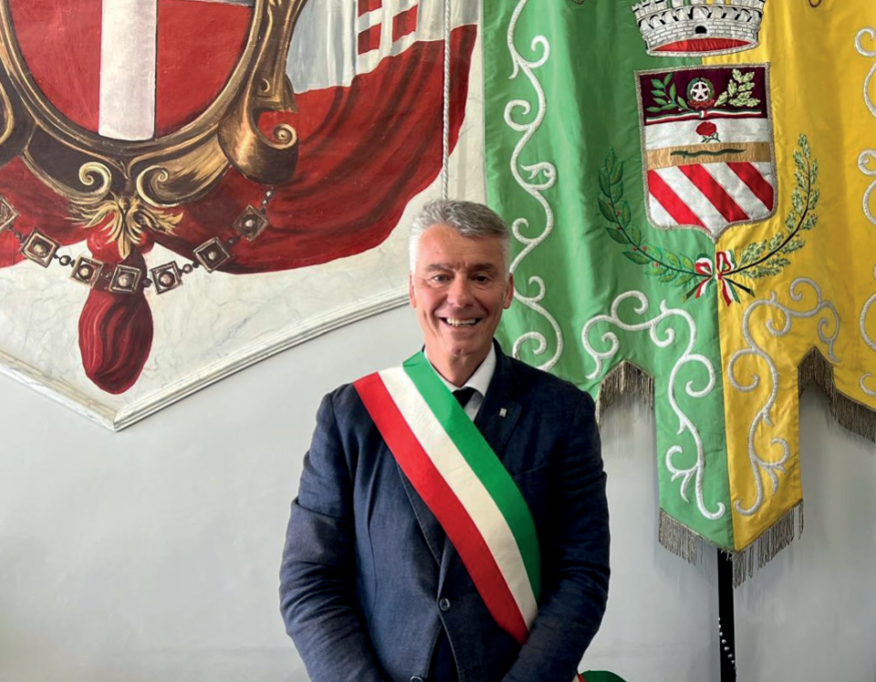 Un uomo sorridente con indosso una fascia è in piedi davanti a striscioni decorativi con simboli araldici, che ricordano una scena del servizio giornalistico sul web di La Sentinella su Roma.